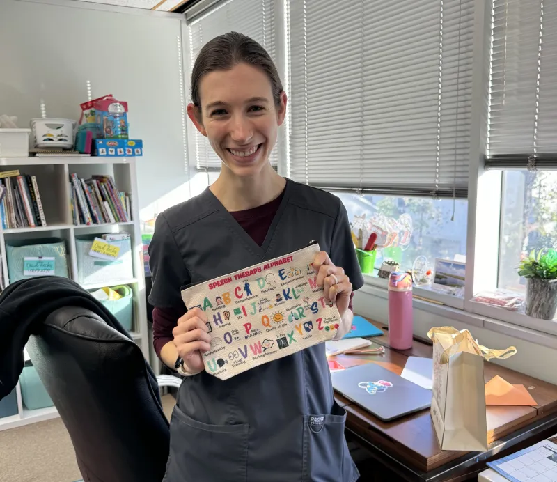 Smiling speech therapist holding a speech therapy alphabet poster in her office